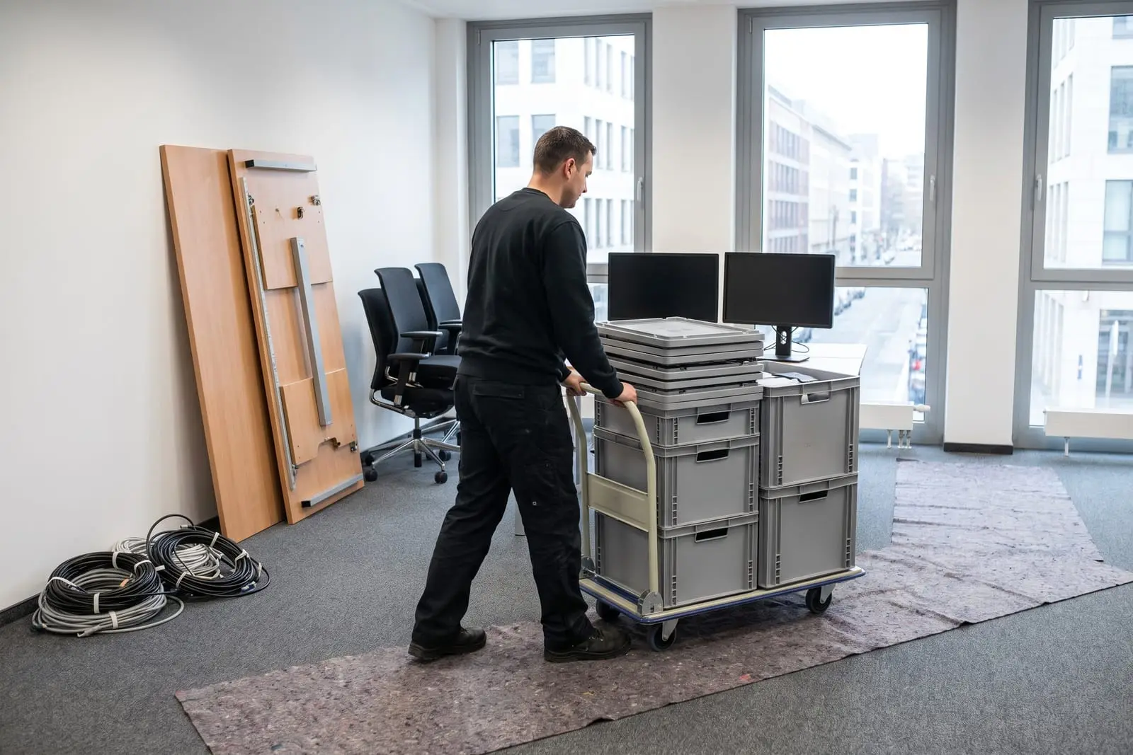 Transportkisten auf einem Rollwagen und bereits abgebaute Schreibtische in einem Büro mit großen Fenstern