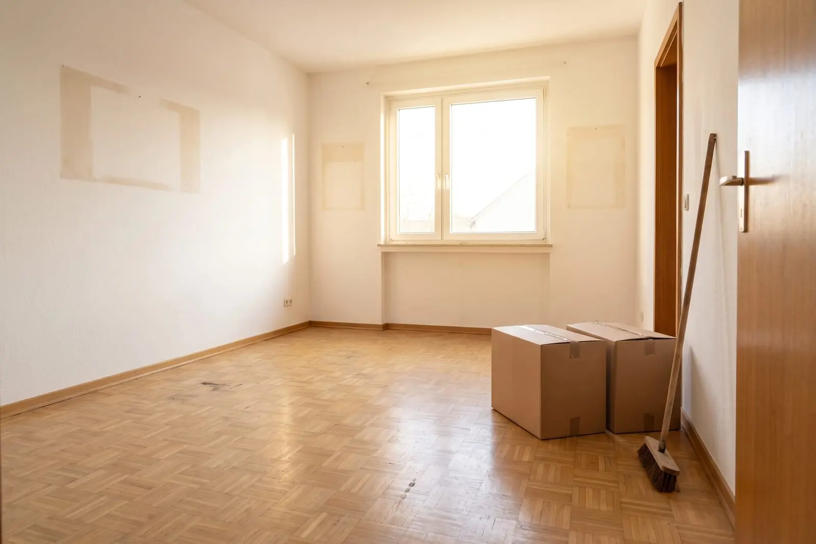 Leergeräumtes Zimmer mit Parkett, zwei Umzugskartons an der Tür und ein Besen an der Wand